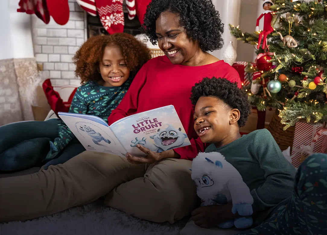 A mother, a girl, and a boy sitting in a living room decorated for Christmas. The mother is holding the Little Grunt storybook while the children read over her shoulder. The boy is holding a Little Grunt plush in his arms.