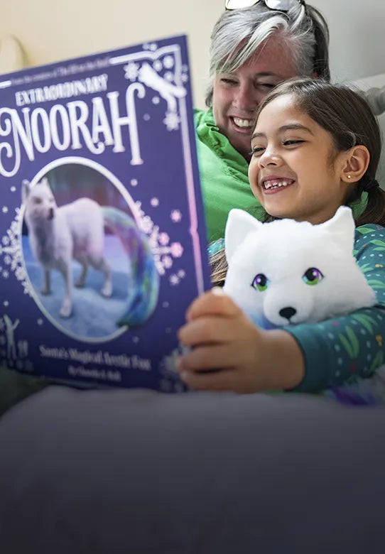 Woman and girl sitting in bed reading the Noorah storybook. Under the girl's arm is the Noorah plush.