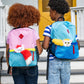 A girl with the red Bogie™ Plush in her colorful backpack walks with a boy who has the yellow Bogie™ Plush in his colorful backpack as they approach their home's front porch