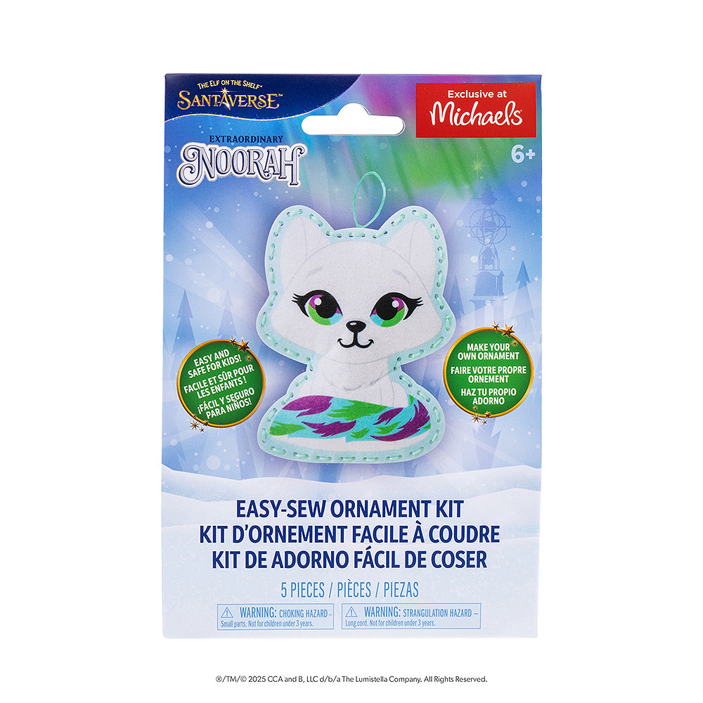 A front view of the packaging of the Noorah® Easy-Sew Ornament Kit. The packaging has the finished product, The Elf on the Shelf and Santaverse logos and other details. The packaging also has an "Exclusive at Michaels" label on it in the top right-hand corner. The background is a vector background commonly associated with the Santaverse brand, featuring conifer trees, light bursts, snowflakes, the Northern Lights, and a snow-covered landscape.
