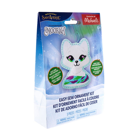 A front view of the packaging of the Noorah® Easy-Sew Ornament Kit, which is slightly turned to the right. The packaging has the finished product, The Elf on the Shelf and Santaverse logos and other details. The packaging also has an "Exclusive at Michaels" label on it in the top right-hand corner. The background is a vector background commonly associated with the Santaverse brand, featuring conifer trees, light bursts, snowflakes, the Northern Lights, and a snow-covered landscape.