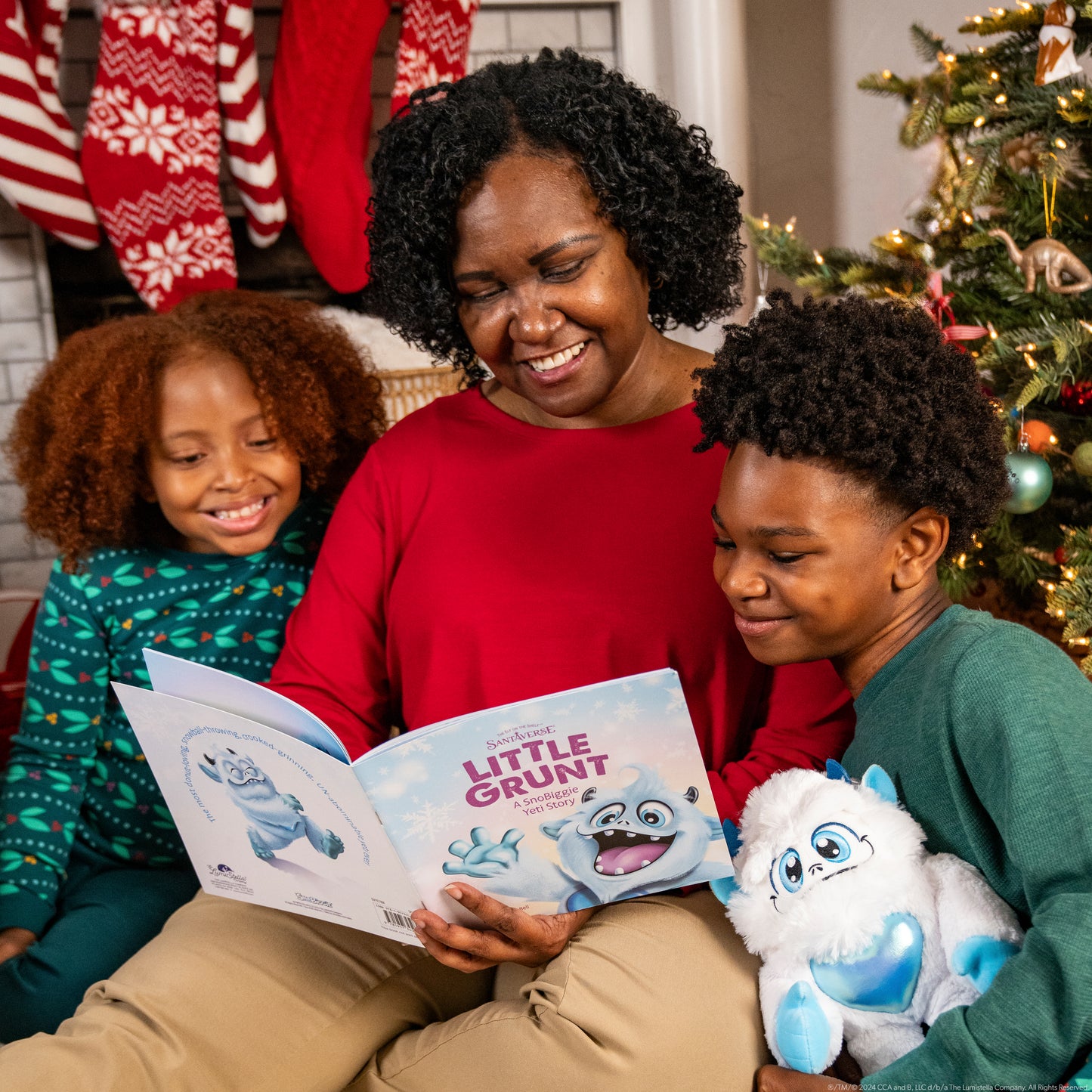 A mother and her children reading the Little Grunt Book, the little boy is holding the Little Grunt Plush, they are sitting in front of their Christmas tree and you can see their fireplace and stockings hanging in the background