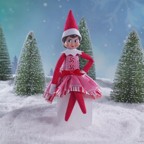 A girl Scout Elf® sitting in a snowy forest donning the Claus Couture Collection® Pink Peppermint Party Dress, spinning around in this product video to give customers a 360 degree view of the product