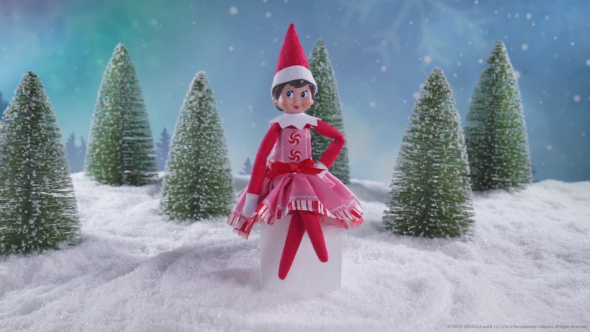 A girl Scout Elf® sitting in a snowy forest donning the Claus Couture Collection® Pink Peppermint Party Dress, spinning around in this product video to give customers a 360 degree view of the product