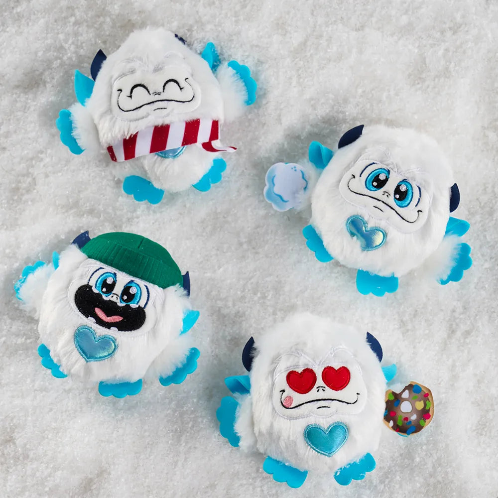 SnoBiggie™ Yeti SnoBall Surprise plush with beanie, SnoBiggie™ Yeti SnoBall Surprise plush with donut and heart eyes, SnoBiggie™ Yeti SnoBall Surprise plush with candy cane pattern scarf, and SnoBiggie™ Yeti SnoBall Surprise plush with snowball in hand all laying on a patch of snow